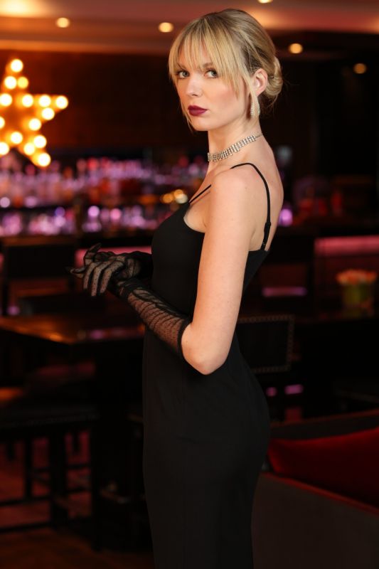Robe de soirée noire ANAE, ROBE MILLIE, parfaite pour les événements chic, mise en valeur par un éclairage ambiant chaud.