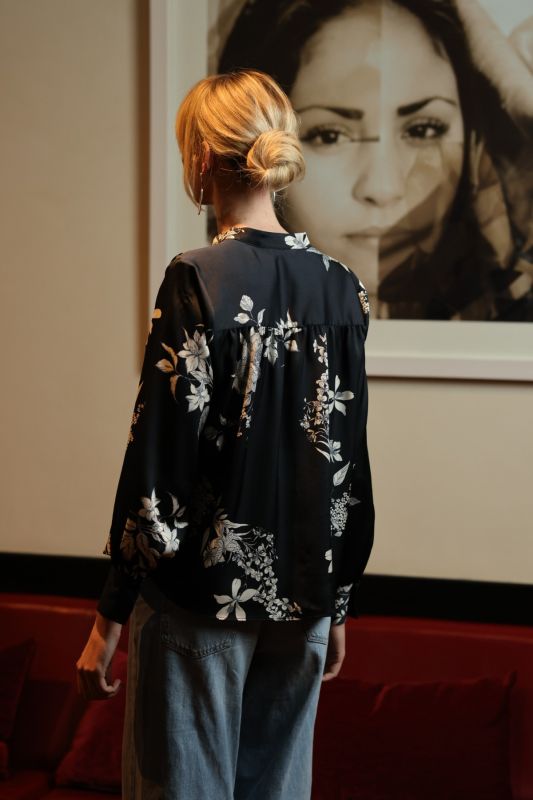 Une femme incarne le charme ANAE dans une blouse noire à imprimé botanique, combinée à un jean, dans un décor artistique.
