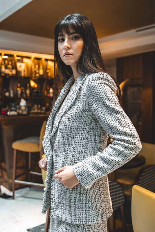 Femme portant une veste en tweed ANAE grise et blanche, style sophistiqué avec un col en frange, dans un cadre de bar.
