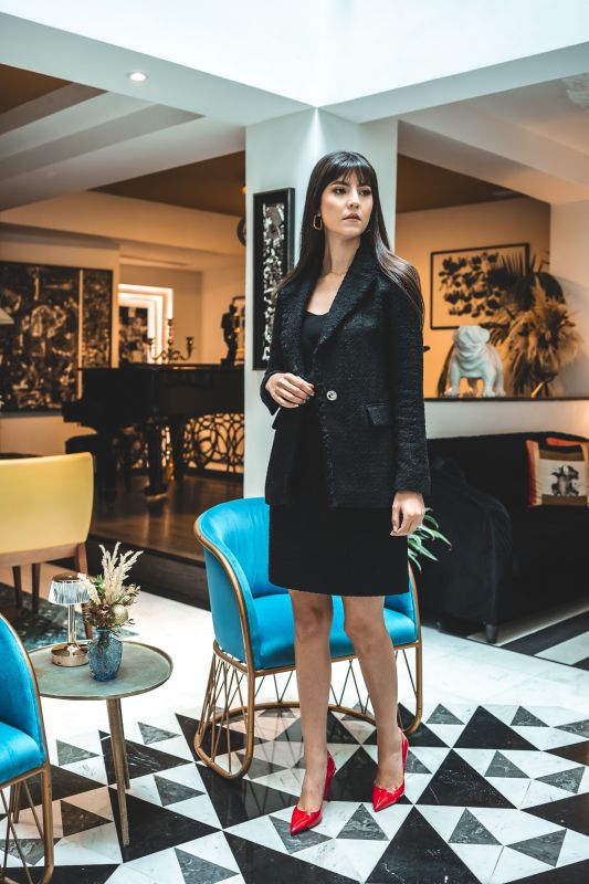 Femme chic assise en veste tweed noire ALISIA TWEED 24 d'ANAE, combinée avec des escarpins rouges pour un contraste audacieux.