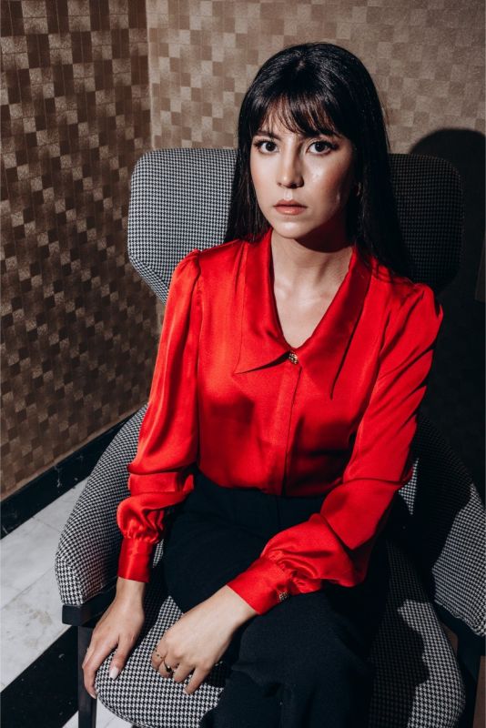 Femme assise en chemise rouge Hardenberg ANAE, avec un regard captivant, évoquant une élégance décontractée.