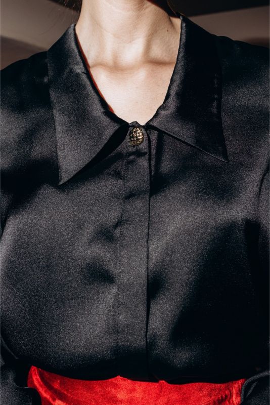 Gros plan sur une chemise noire ANAE avec bouton bijou unique, contrastant superbement avec une jupe rouge satinée.