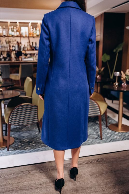 Femme élégante avec manteau bleu Chaouen d'ANAE, ajustant sa manche, dans un intérieur chic et contemporain.