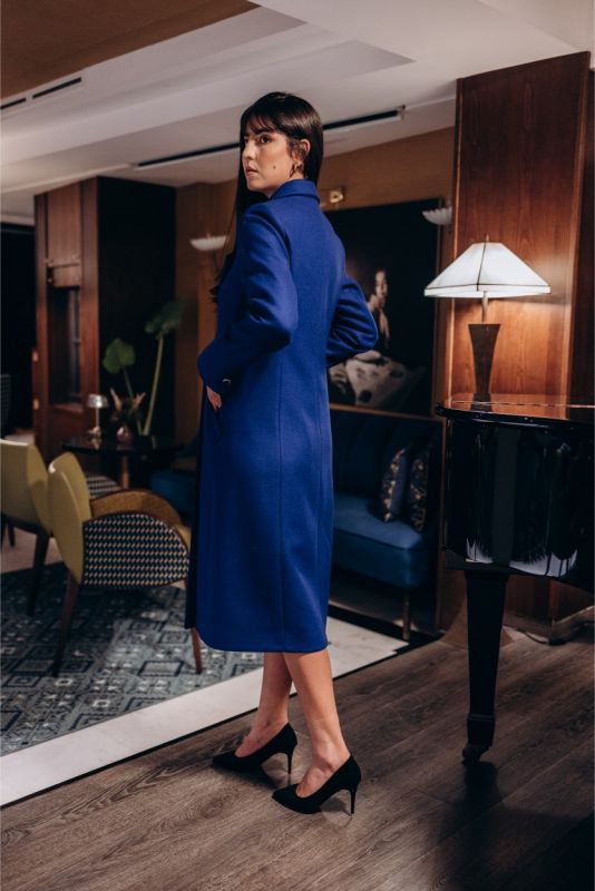 Femme élégante avec manteau bleu Chaouen d'ANAE, ajustant sa manche, dans un intérieur chic et contemporain.