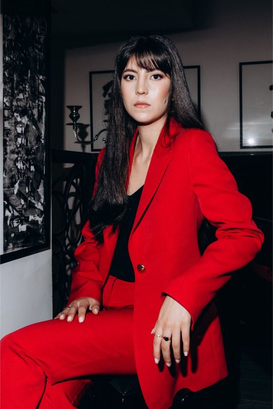 Femme élégante en tailleur pantalon rouge vif de la collection ANAE, assise dans une pièce au style contemporain.