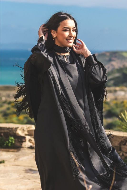 Femme souriante en caftan Anissa noir, détails de franges, sur fond marin d'ANAE.