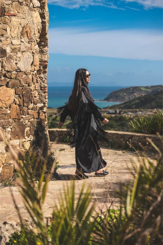 Caftan Anissa noir d'ANAE avec franges, devant un mur de pierre pour un look Ramadan.