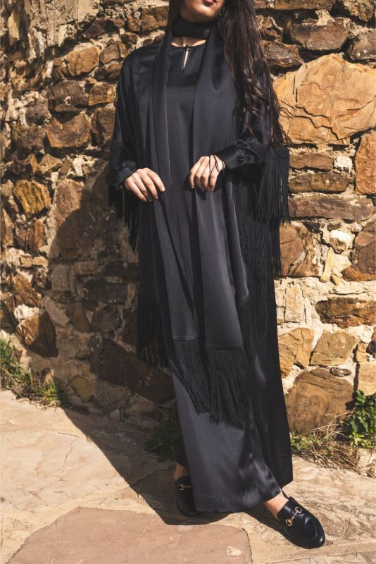 Caftan Anissa noir d'ANAE avec franges, devant un mur de pierre pour un look Ramadan.