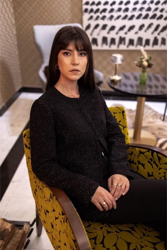 Femme assise en veste Margaret noire, ambiance de luxe contemporain avec une touche de brillance.