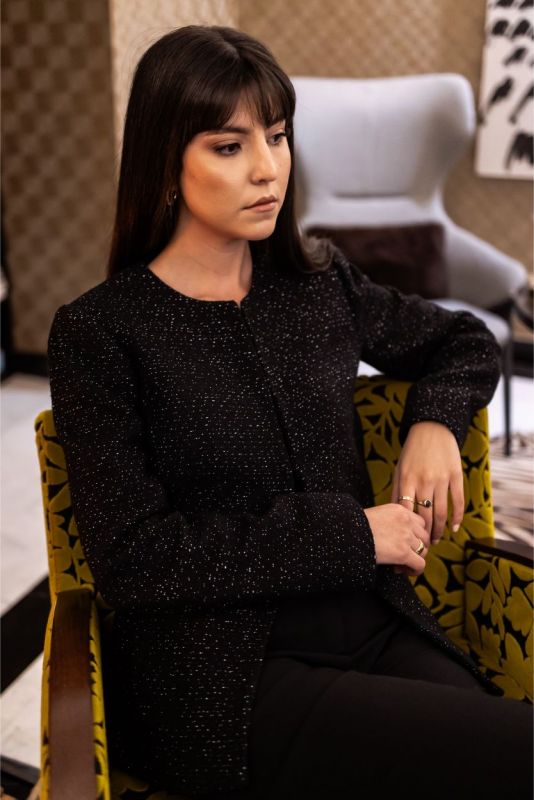 Femme assise en veste Margaret noire, ambiance de luxe contemporain avec une touche de brillance.