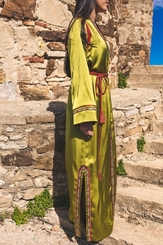 Caftan FAIROUZ d'ANAE en vert olive, détails de couture rouge et dorée, posant sur des marches en pierre.