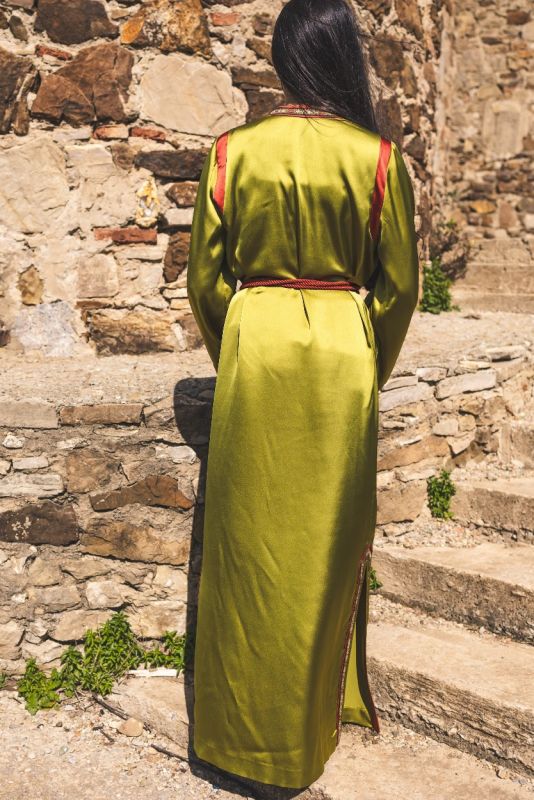 Caftan FAIROUZ d'ANAE en vert olive, détails de couture rouge et dorée, posant sur des marches en pierre.