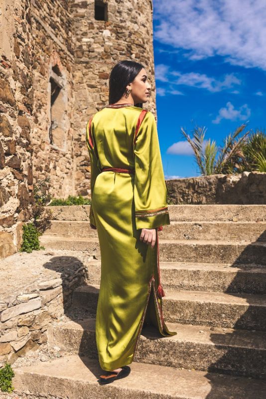 Femme vêtue du caftan FAIROUZ d'ANAE reposant sur des marches antiques, mêlant tradition et modernité.