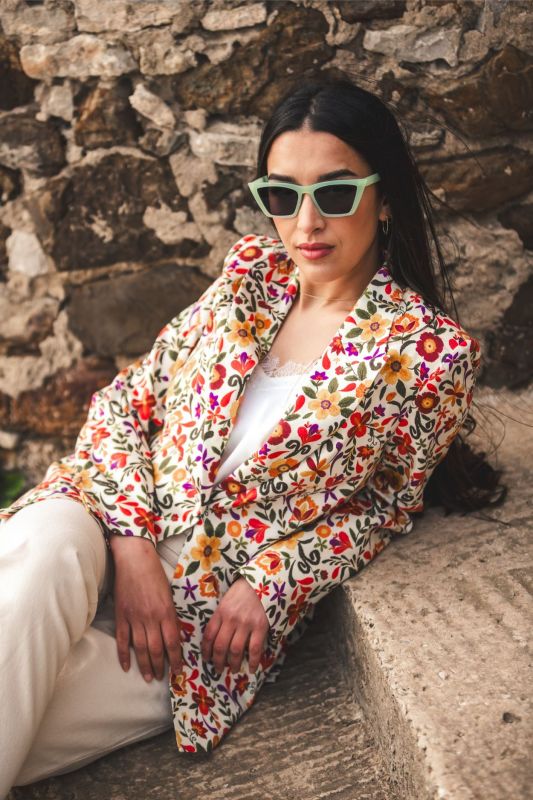 Veste JEANNA fleurie d'ANAE, style décontracté chic avec lunettes de soleil vertes, devant un mur de pierres.