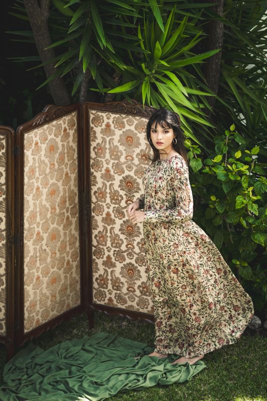 Robe PALM longue d'ANAE dans un jardin, présentant un motif floral sur fond crème et une coupe fluide.