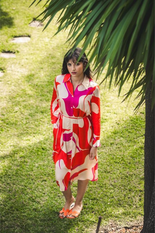 Femme posant sous les palmiers en robe 'Amalia' mi-longue, motif rouge et rose, et sandales orange vives pour un style estival.