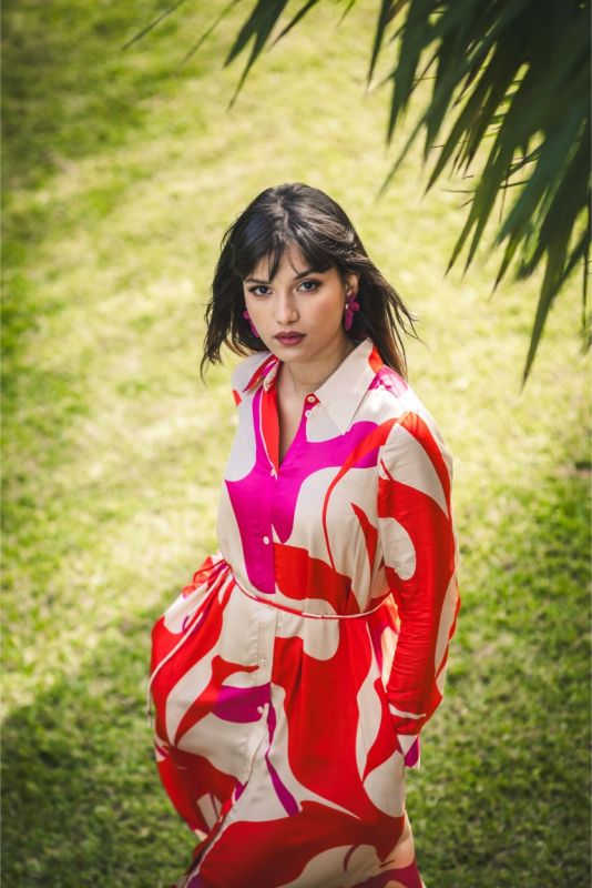 Femme posant sous les palmiers en robe 'Amalia' mi-longue, motif rouge et rose, et sandales orange vives pour un style estival.