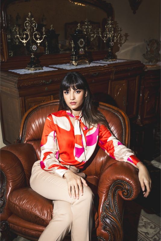 Femme portant une blouse rouge à motifs abstraits et un pantalon beige, vue de face.