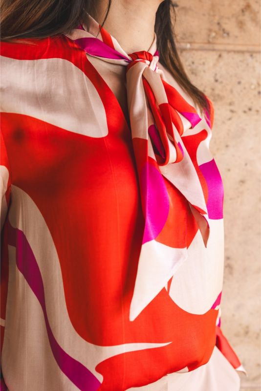 Gros plan sur la blouse 'Carina' d'ANAE avec détail de noeud, manches bouffantes et imprimé rouge vif et rose sur fond beige.