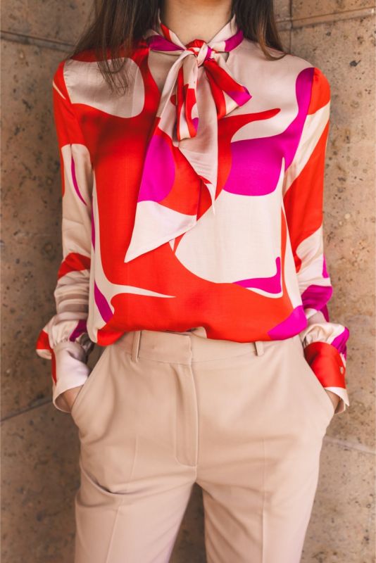 Femme assise dans un fauteuil en cuir, portant une blouse rouge à motifs abstraits et un pantalon beige.