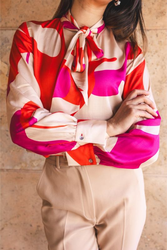Femme assise dans un fauteuil en cuir, portant une blouse rouge à motifs abstraits et un pantalon beige.