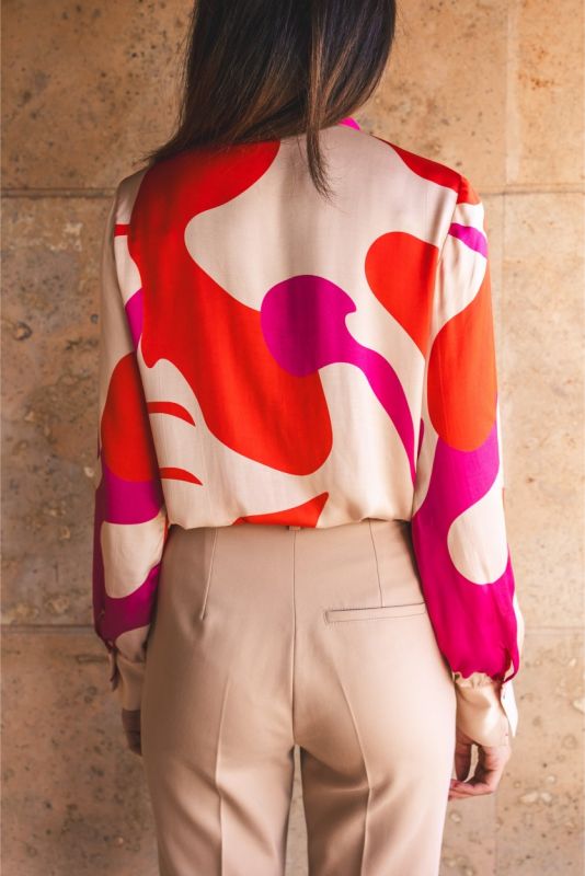 Femme assise dans un fauteuil en cuir, portant une blouse rouge à motifs abstraits et un pantalon beige.