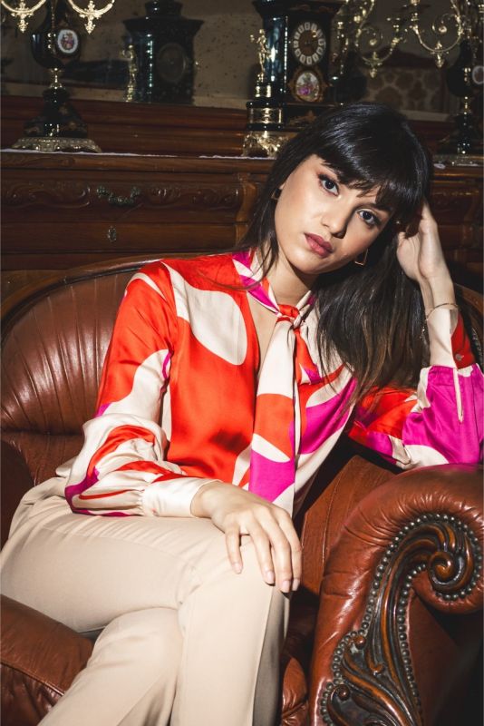 Femme portant une blouse rouge à motifs abstraits et un pantalon beige, vue de face.