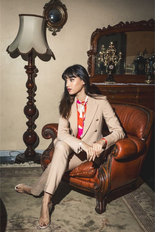 Jeune femme en veste 'Frida' beige à double boutonnage et détails boutons dorés, assise élégamment sur un fauteuil en cuir.