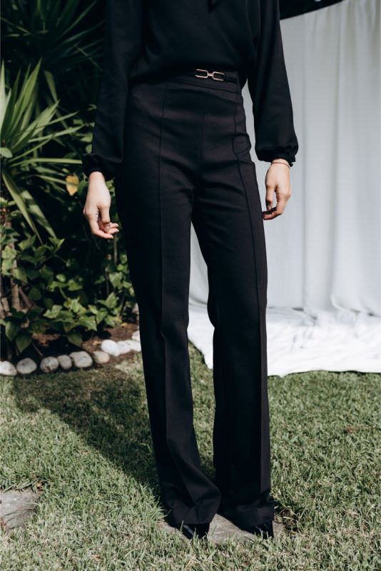 Femme élégante en pantalon noir évasé et blouse noire avec nœud lavallière, ambiance extérieure naturelle.
