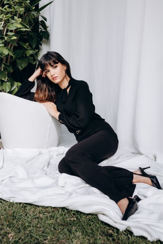 Femme élégante en pantalon noir évasé et blouse noire avec nœud lavallière, ambiance extérieure naturelle.