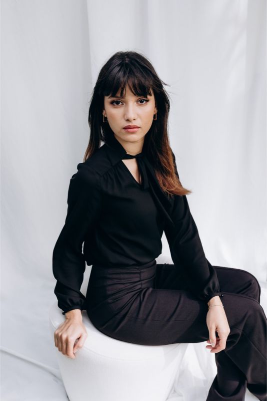 Femme élégante en blouse noire 'Belkis' avec nœud au col, s'appuyant sur un sofa blanc, une touche de sophistication ANAE.