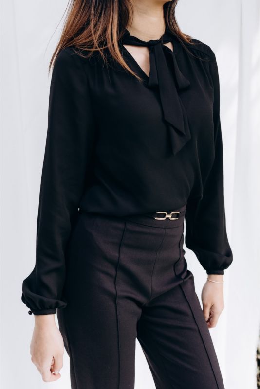 Portrait assis d'une femme portant la blouse noire 'Belkis', style épuré avec nœud délicat, reflet du chic urbain par ANAE.