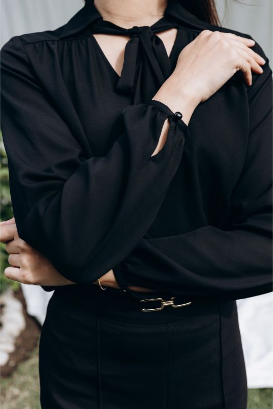 Portrait assis d'une femme portant la blouse noire 'Belkis', style épuré avec nœud délicat, reflet du chic urbain par ANAE.