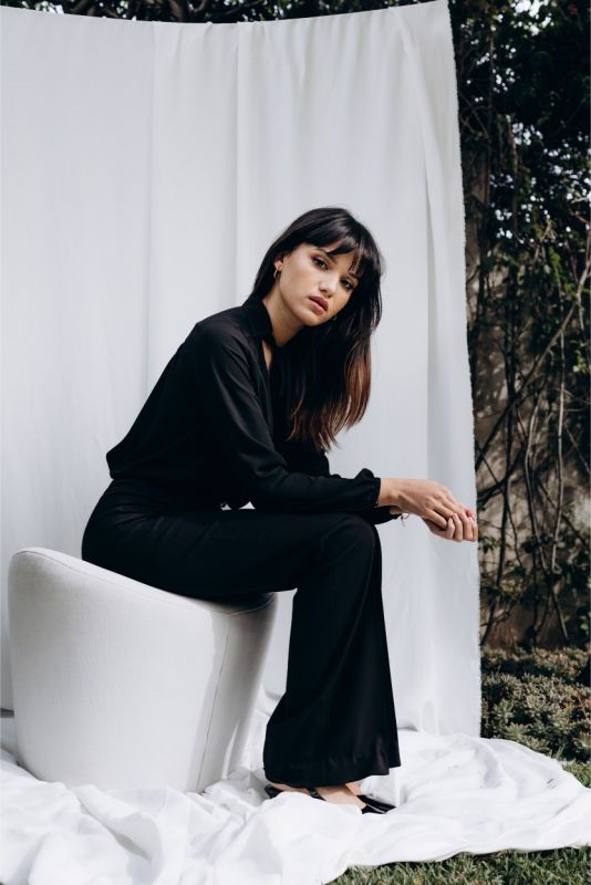 Femme élégante en blouse noire 'Belkis' avec nœud au col, s'appuyant sur un sofa blanc, une touche de sophistication ANAE.