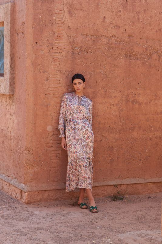 Femme en robe paisley regardant au loin, portant des lunettes de soleil blanches, contre un fond de mur en terre.