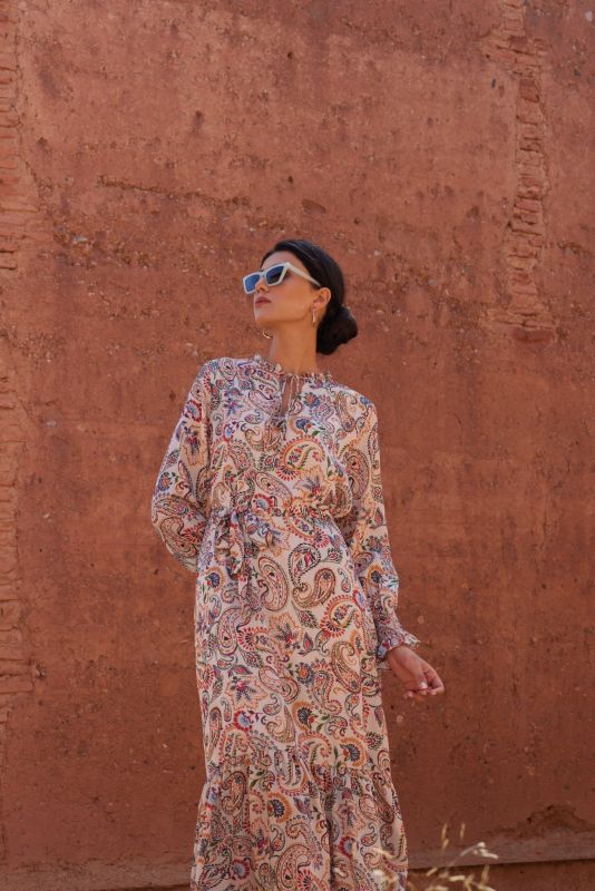 Femme en robe paisley regardant au loin, portant des lunettes de soleil blanches, contre un fond de mur en terre.