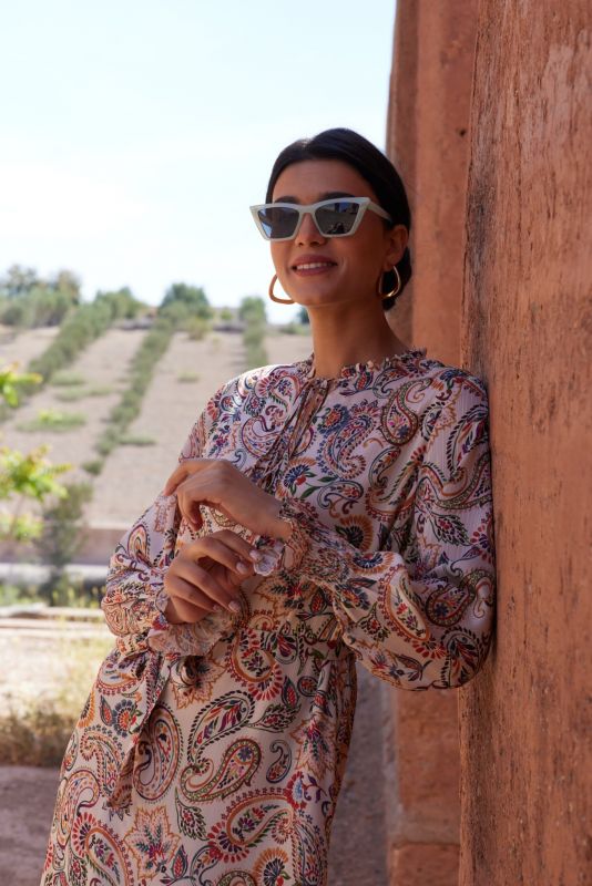Plan rapproché d'une femme en robe beige imprimée, lunettes de soleil blanches, fond de mur en terre cuite.