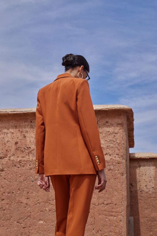 Femme en PANTALON ROSARIO orange et blazer assorti, debout sur un mur de terre cuite, incarnant le chic moderne.