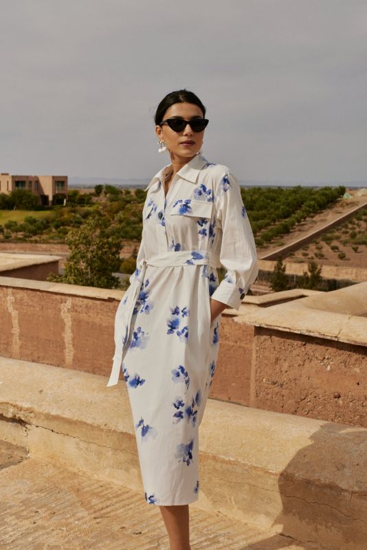 Femme portant une robe chemise blanche à motifs floraux bleus, avec une ceinture nouée, posant devant un paysage montagneux.