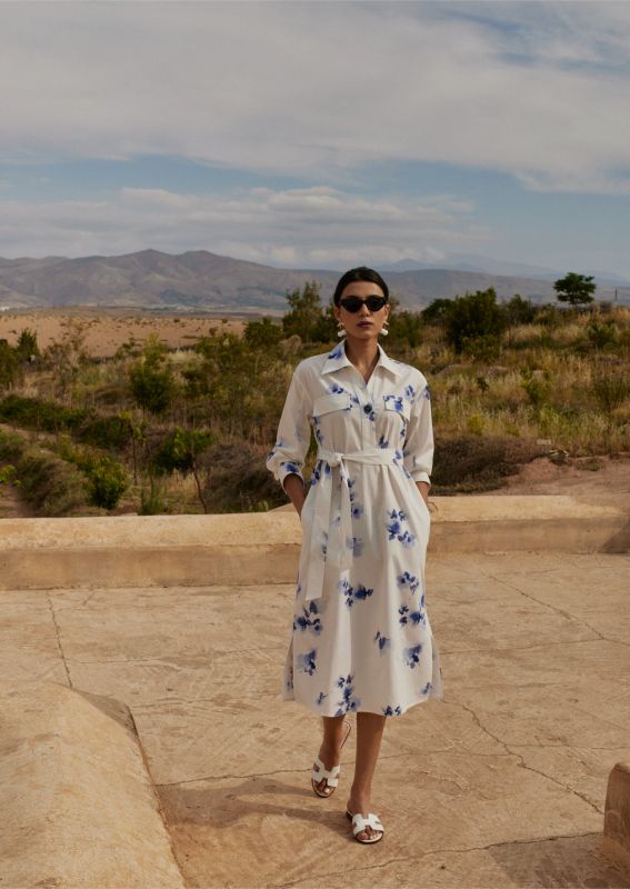 Femme portant une robe chemise blanche à motifs floraux bleus, avec une ceinture nouée, posant devant un paysage montagneux.