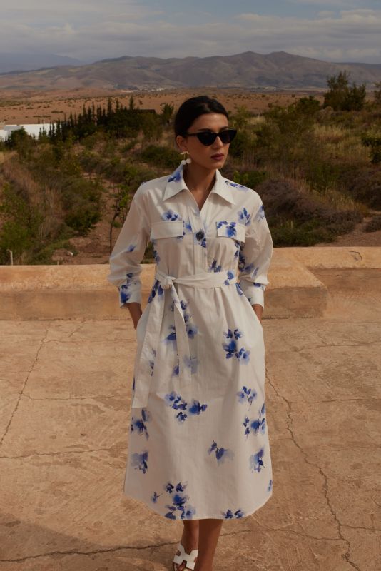 Vue de dos d'une femme en robe chemise blanche à motifs floraux bleus, avec ceinture nouée, devant un paysage naturel.