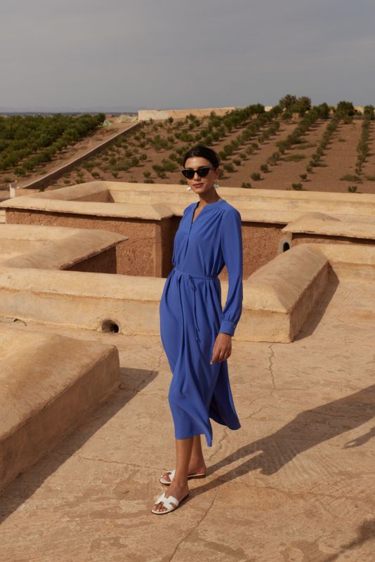 Femme portant une robe mauve avec manches longues et ceinture nouée, posant devant un mur de pierre, regardant au loin.