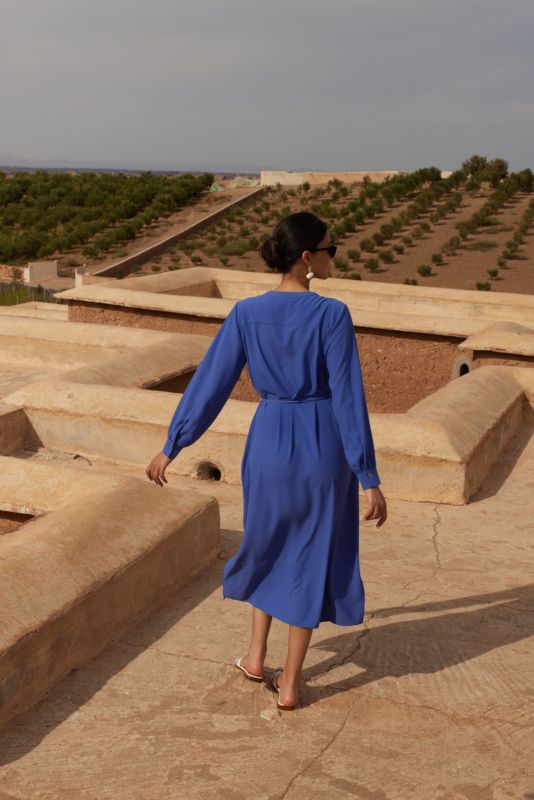 Femme portant une robe mauve avec manches longues et ceinture nouée, posant devant un mur de pierre, regardant au loin.