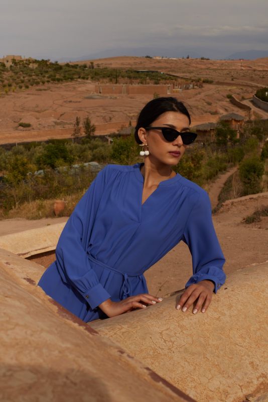 Femme portant une robe mauve avec manches longues et ceinture nouée, posant devant un mur de pierre, regardant au loin.