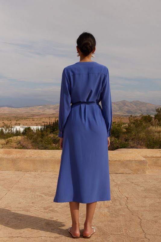 Femme portant une robe mauve avec manches longues et ceinture nouée, posant devant un mur de pierre, regardant au loin.