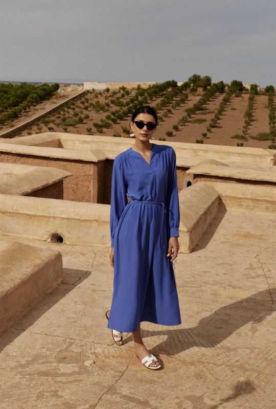 Femme en robe mauve à manches longues et ceinture nouée, marchant sur une terrasse en pierre avec des collines en arrière-plan.
