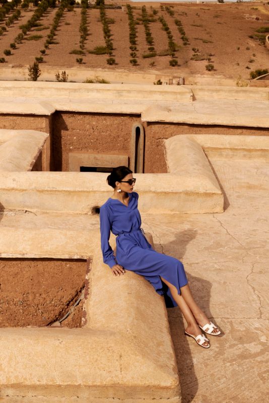 Femme portant une robe mauve avec manches longues, ceinture nouée et lunettes de soleil noires, posant devant un ciel dégagé.