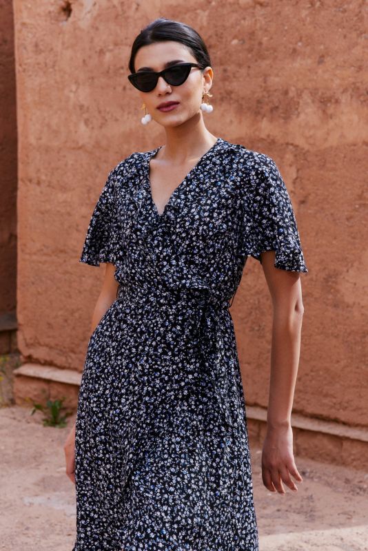 Femme portant une robe noire à motifs floraux blancs, les bras levés, posant entre deux murs en terre cuite.