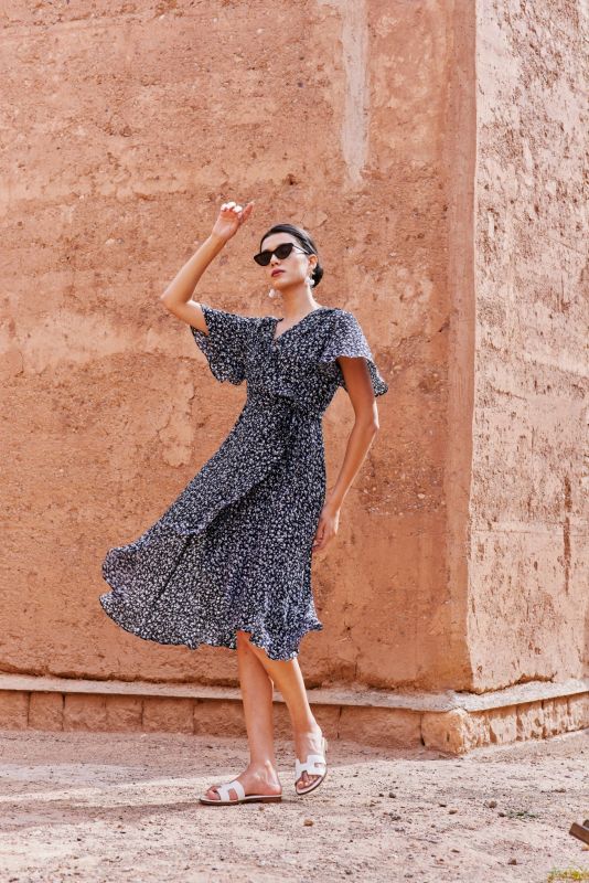 Femme portant une robe noire à motifs floraux blancs, posant contre un mur en terre cuite.
