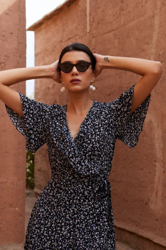 Femme portant une robe noire à motifs floraux blancs, avec des lunettes de soleil et des boucles d'oreilles pendantes.